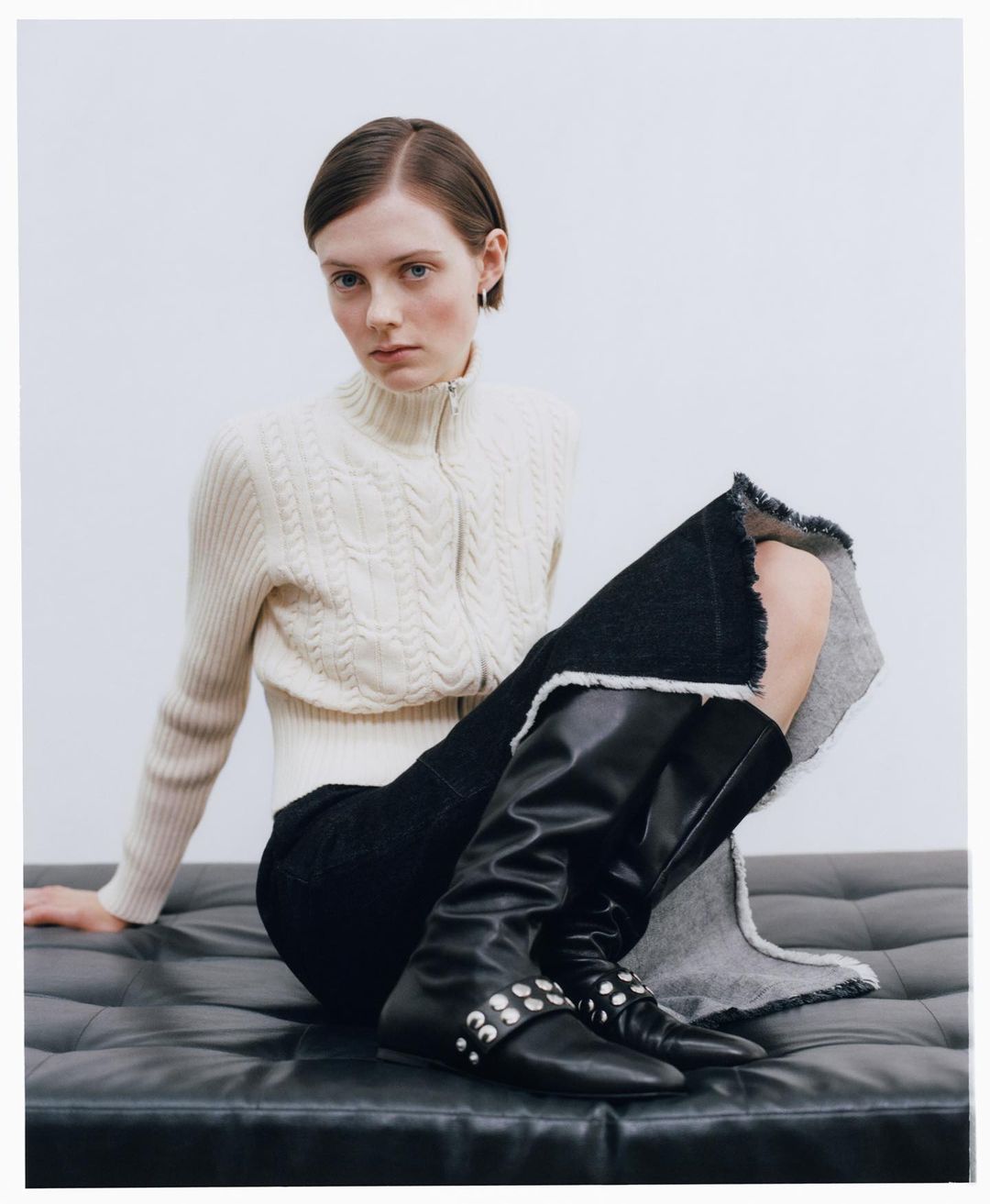 Fashion shot of an adult woman in a sweater and black skirt sitting on a leather bench wearing tall, black studded boots in a studio.