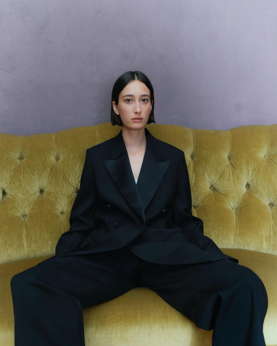 A slender young woman in a black pantsuit sitting on a mustard-colored velvet couch in a portrait-style fashion shot.