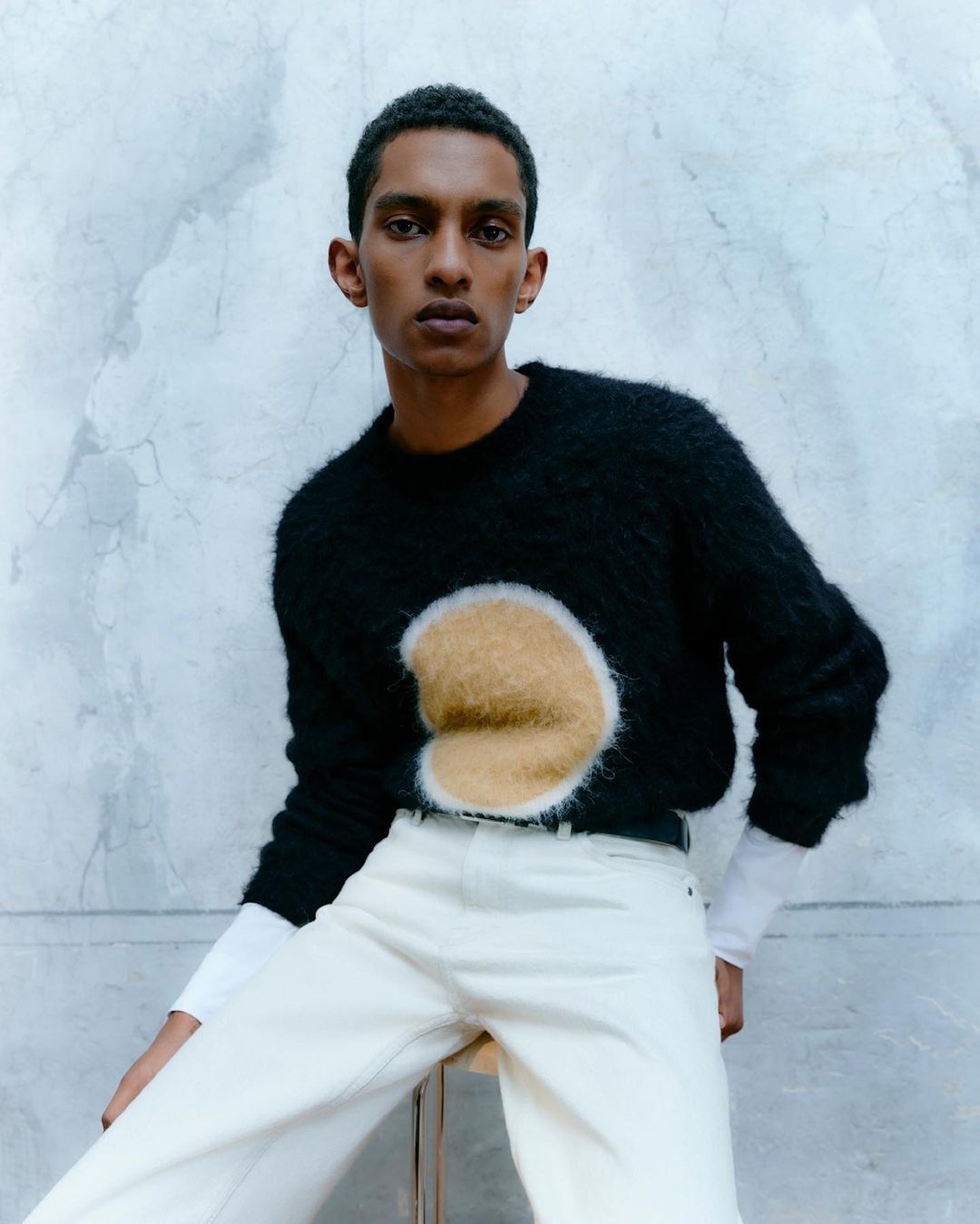 An adult male poses indoors wearing a black sweater with a tan circular design, white jeans, and a black belt in a studio setting.
