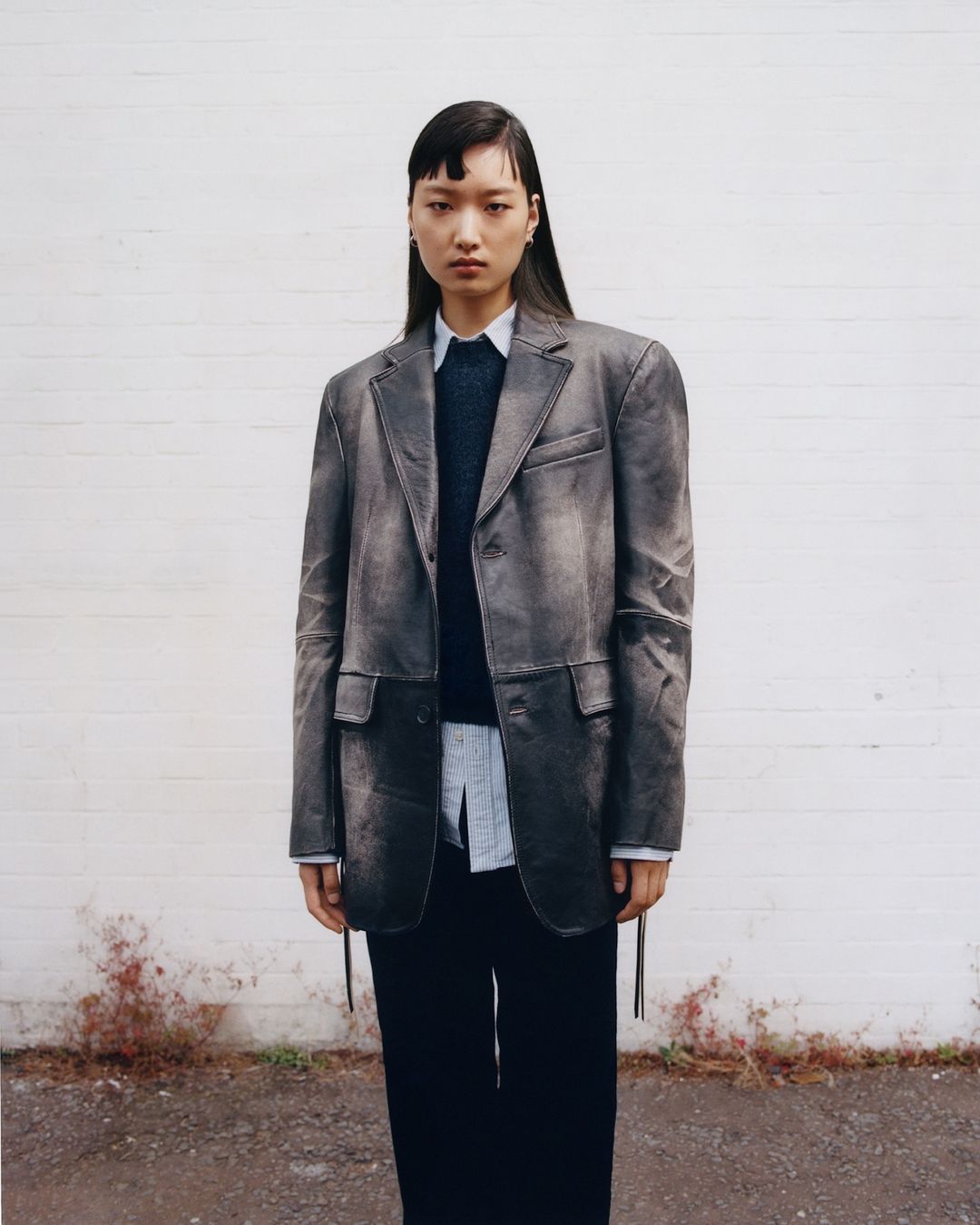 Portrait of an Asian young adult female wearing a grey distressed blazer, black pants, and a white shirt against a white brick wall.
