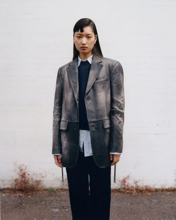 Portrait of an Asian young adult female wearing a grey distressed blazer, black pants, and a white shirt against a white brick wall.