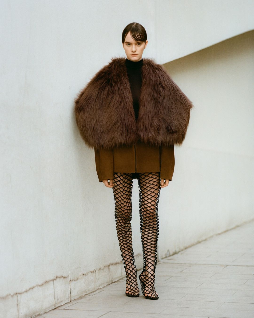 Fashion model wearing a brown fur collar jacket with black top, fishnet tights and heels posing against an off-white colored wall.