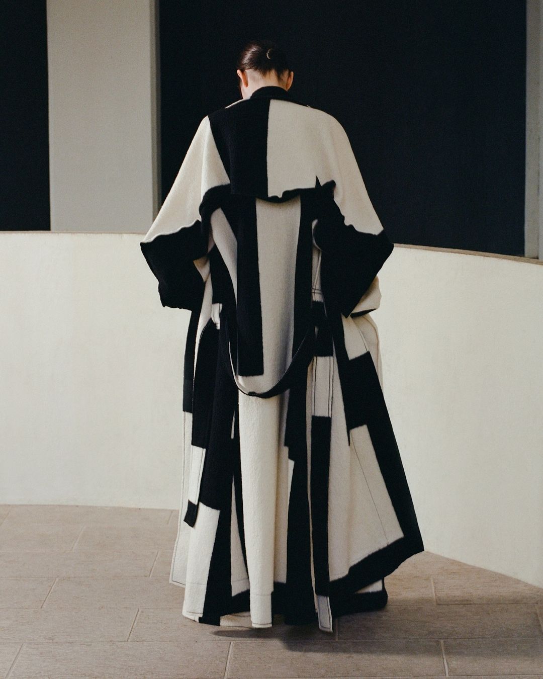 Back view of a person in a black-and-white coat standing on a tiled surface against a dark and light wall indoors.