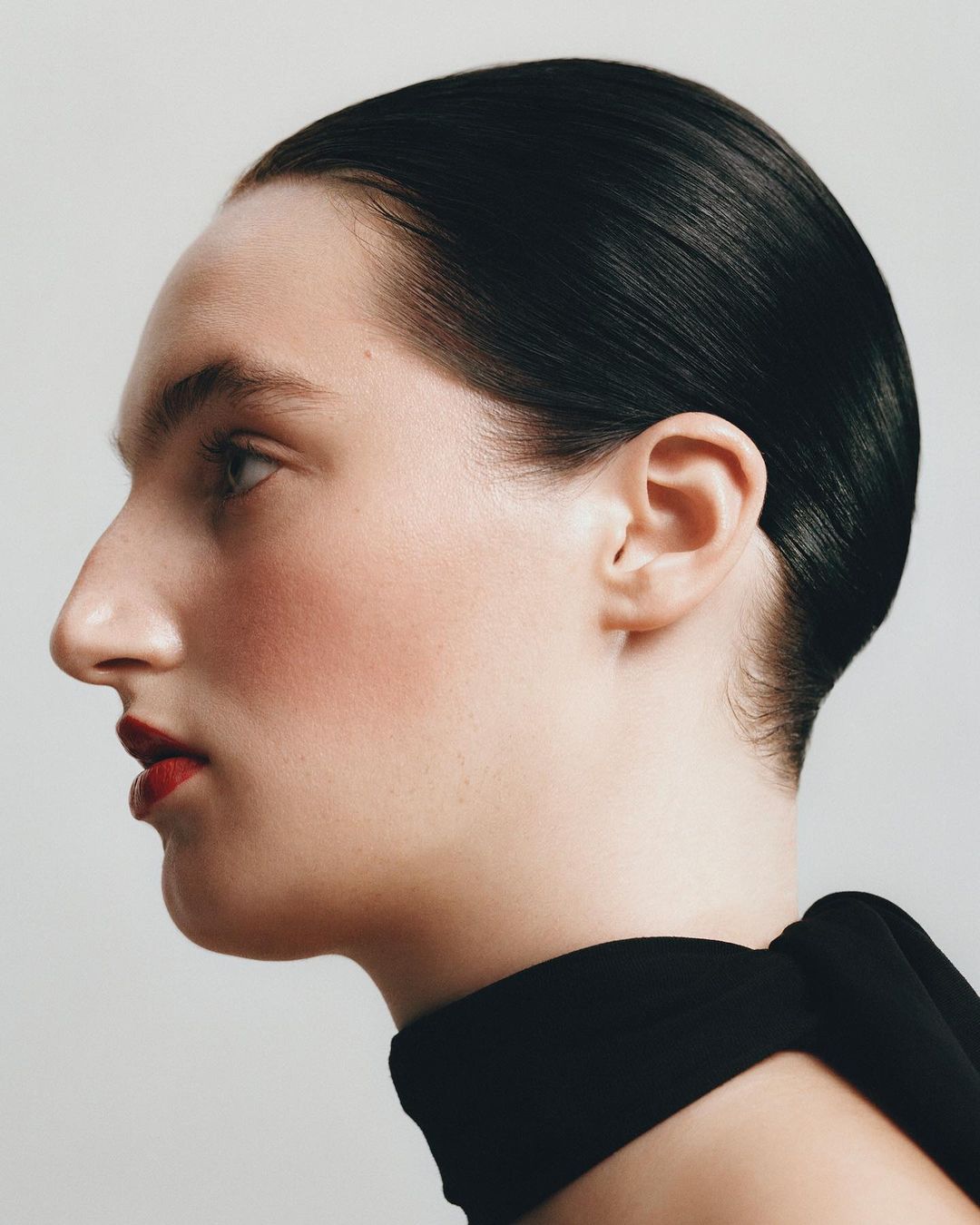 Profile view of a fair-skinned adult female with slicked-back dark hair, red lipstick, and a black garment against a light grey background.
