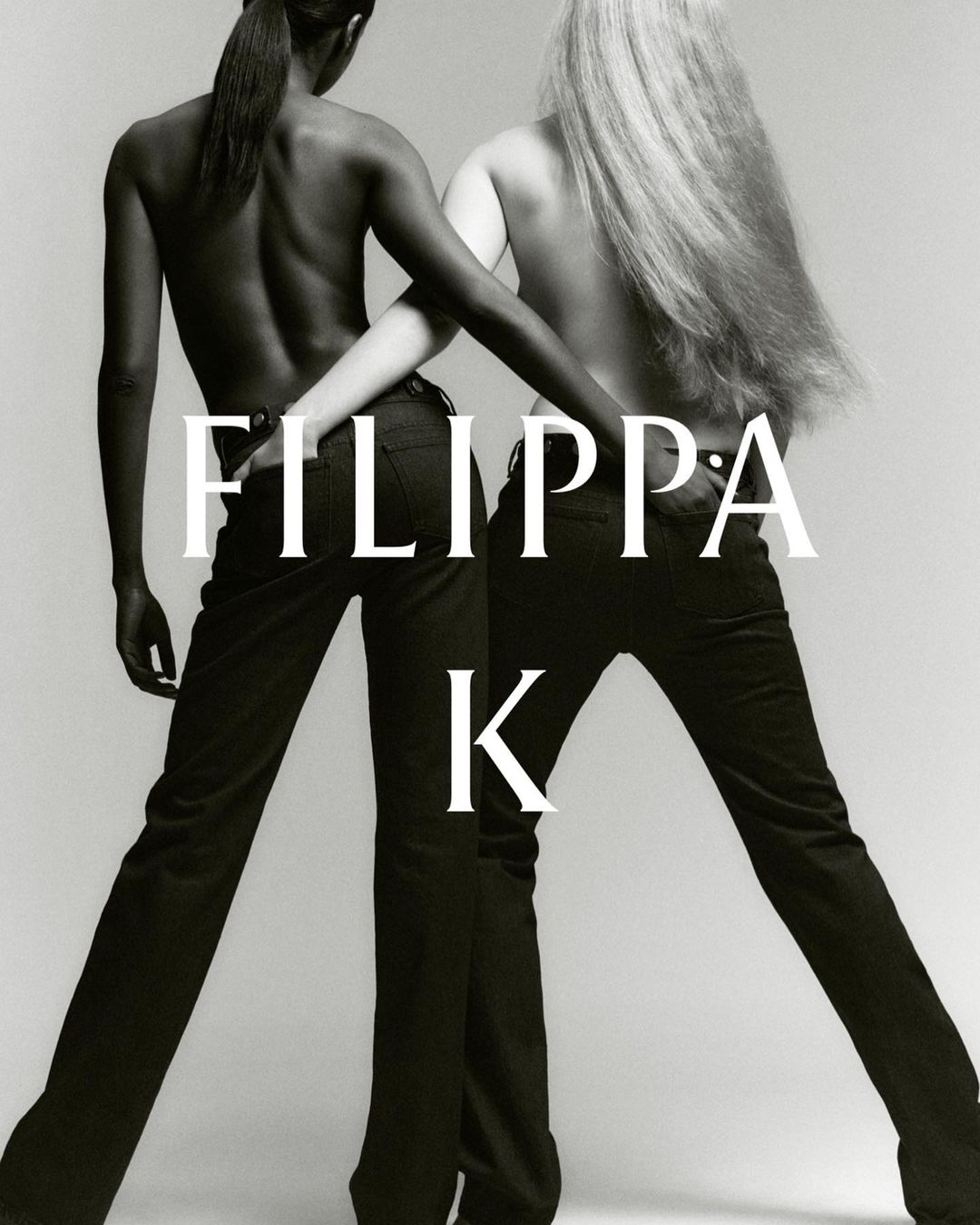 Two fashion models pose with arms linked wearing black trousers against a pale background in a studio portrait for a clothing advertisement.