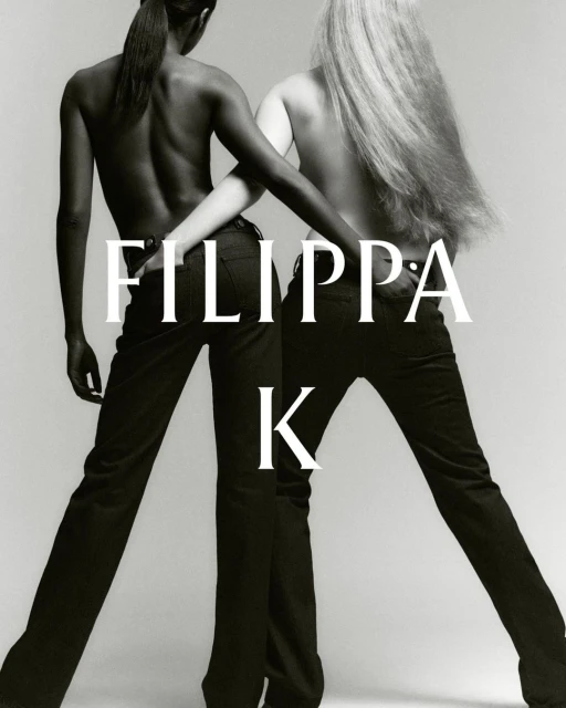 Two fashion models pose with arms linked wearing black trousers against a pale background in a studio portrait for a clothing advertisement.