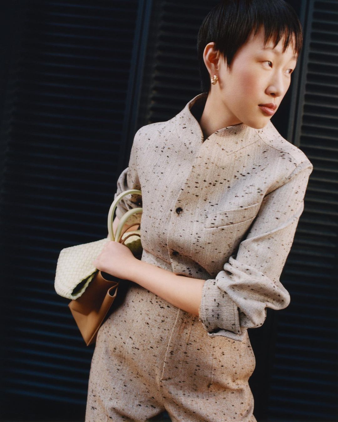 Elegant woman in speckled beige jumpsuit holds a light-colored tote bag against a dark louvered background, exuding a high-fashion editorial aesthetic.