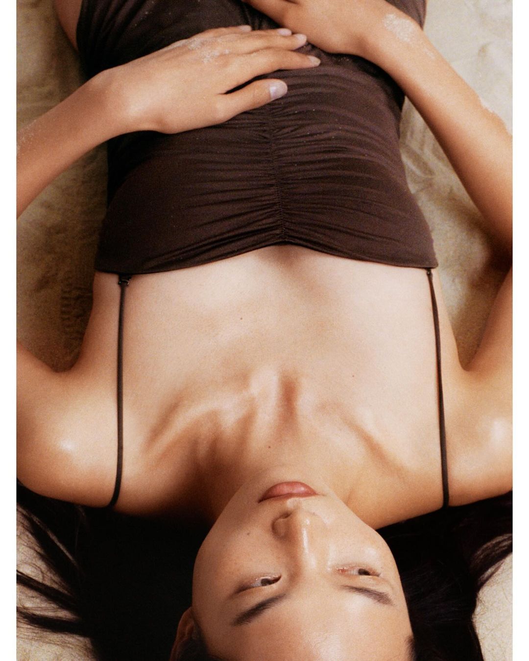 Woman lying down wearing brown top. Overhead shot of female model with dark hair lying on tan colored cloth looking at camera.