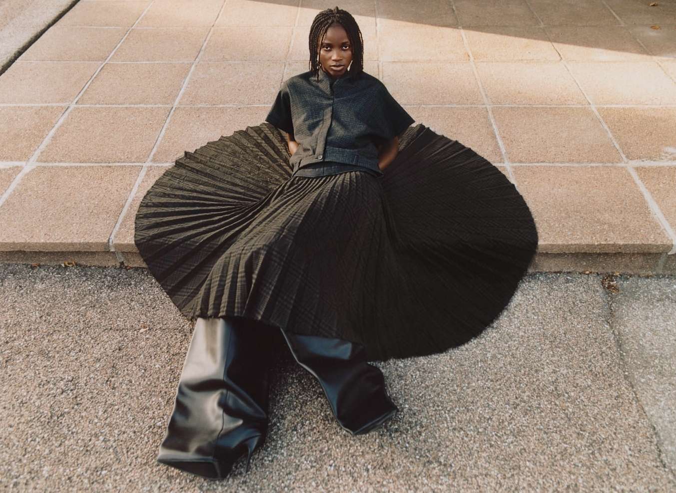 Fashion shot of a stylish woman in a dark top, pleated skirt, and high leather boots, posing on a patterned tile surface outdoors.