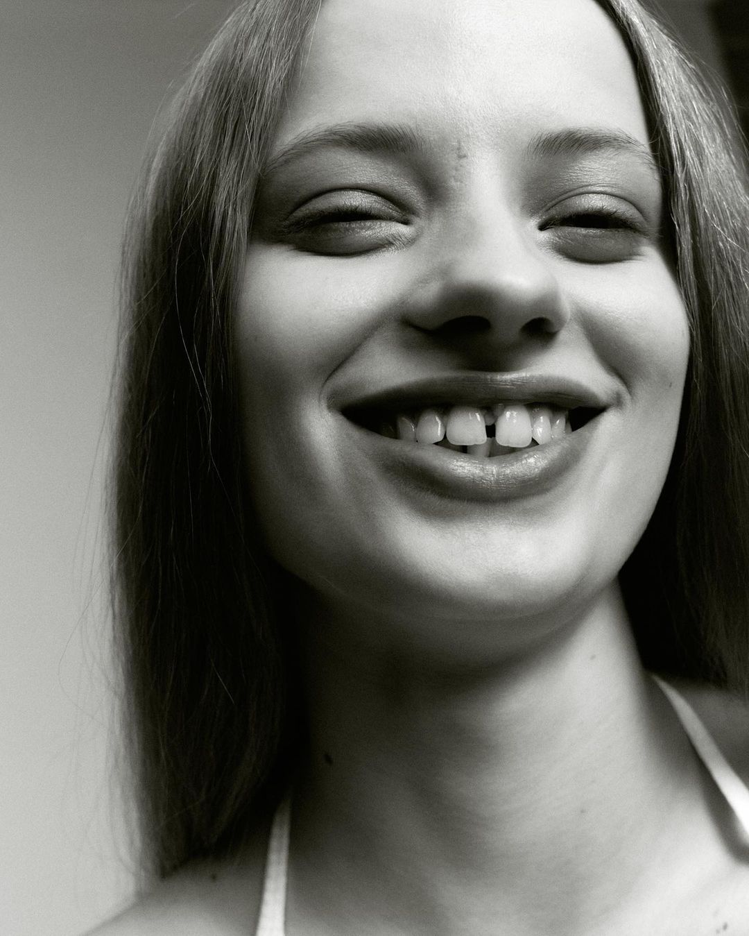 Close-up portrait of a happy, fair-skinned young woman with a gap between her front teeth, smiling at the camera against a neutral background.