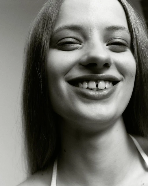 Close-up portrait of a happy, fair-skinned young woman with a gap between her front teeth, smiling at the camera against a neutral background.