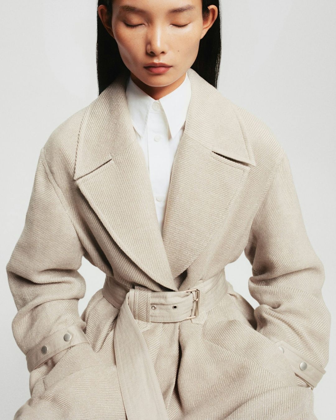 A woman wearing a beige belted coat in a studio setting with a soft focus. The model's eyes are closed, creating a serene and elegant mood.