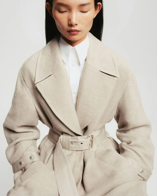 A woman wearing a beige belted coat in a studio setting with a soft focus. The model's eyes are closed, creating a serene and elegant mood.