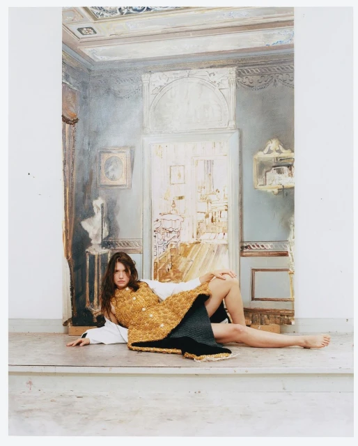 Woman in gold woven vest lying in front of a painted backdrop depicting an interior architectural design in muted colors, studio setting.