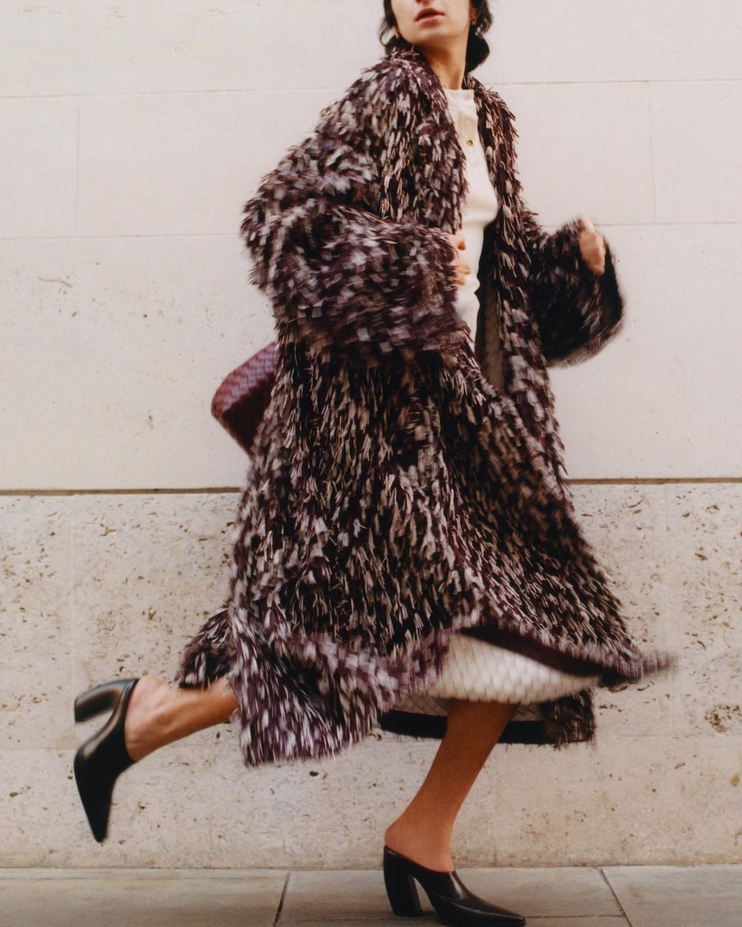 Woman in a fur coat and black heeled mules walking along a city street. She carries a brown woven handbag. The background is a beige wall.