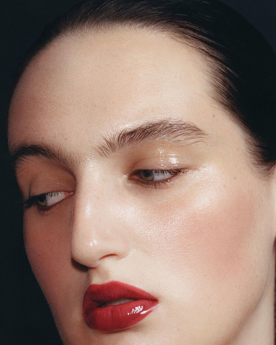 Close-up portrait of an adult woman with glossy red lips and dewy skin, posing against a dark background in soft studio lighting.