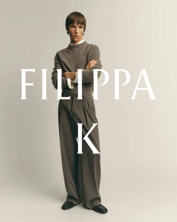 Model with arms crossed wearing a brown knit sweater and trousers against a pale grey background