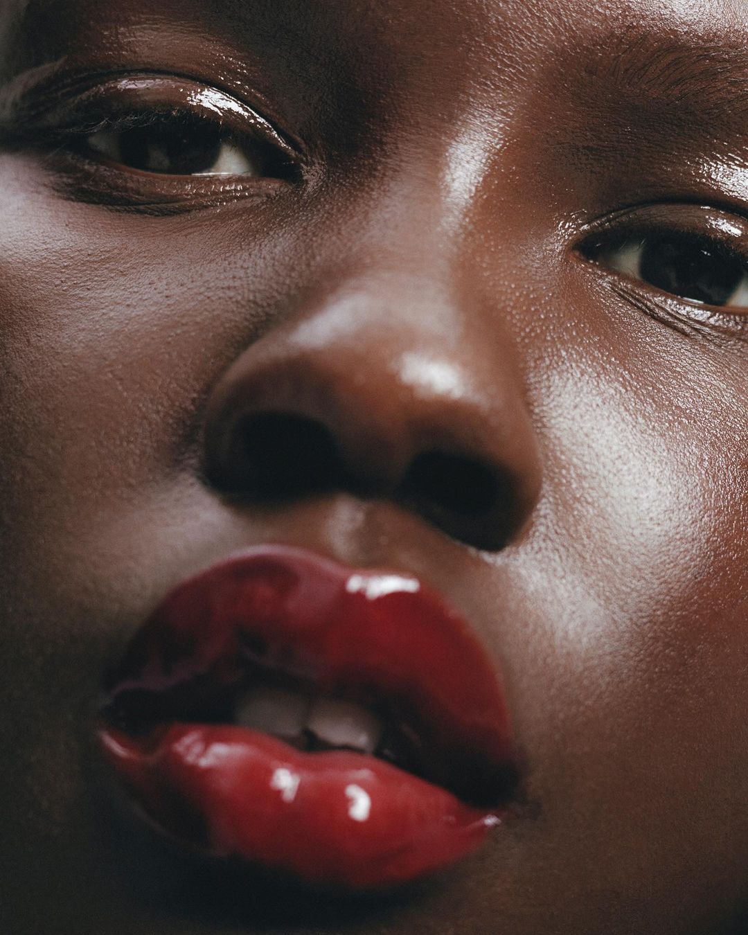 Close-up portrait of a woman with brown skin and glossy red lips, focusing on her facial features with wet, shimmering skin.