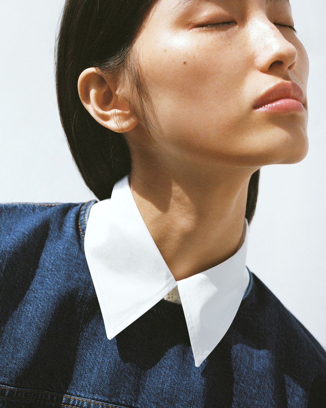 Close-up portrait of a young woman with closed eyes and flawless skin, wearing a denim top with a crisp white collar, against a bright background.
