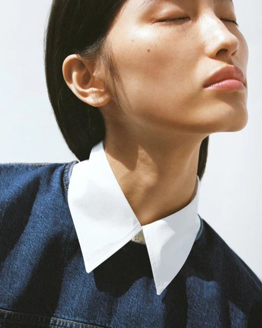 Close-up portrait of a young woman with closed eyes and flawless skin, wearing a denim top with a crisp white collar, against a bright background.
