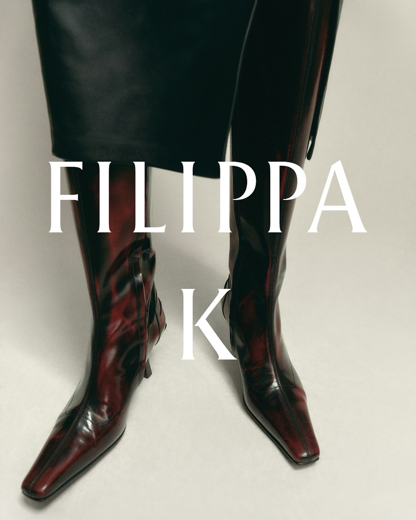 Close-up studio shot of burgundy knee-high leather boots worn with a black skirt, featuring the 'Filippa K' logo.