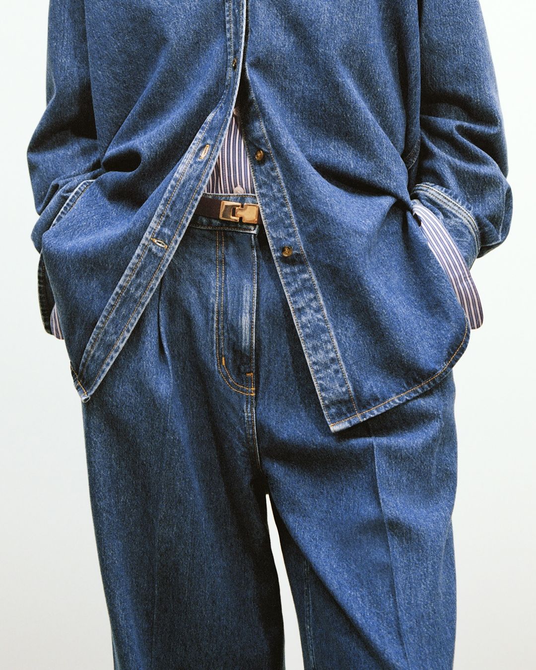 Denim fashion ensemble: a person wears a blue denim shirt, striped shirt, brown leather belt and denim pants against a white background.