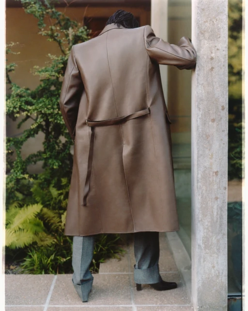Back view of an adult wearing a long brown coat over gray pants and black shoes, leaning on a concrete column outdoors amid greenery.