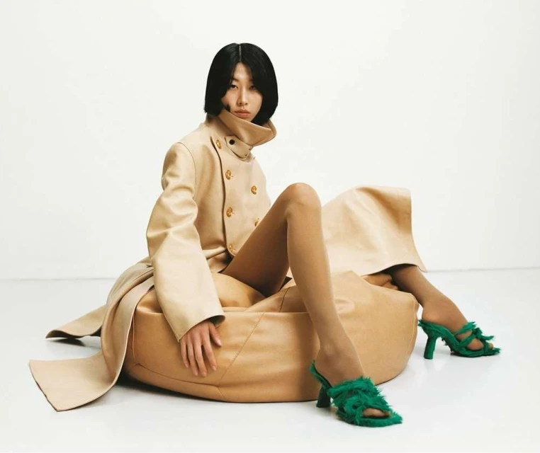 Fashion shot of an Asian model wearing a tan double-breasted coat and green heeled sandals, sitting on a tan leather pouf on a white background.