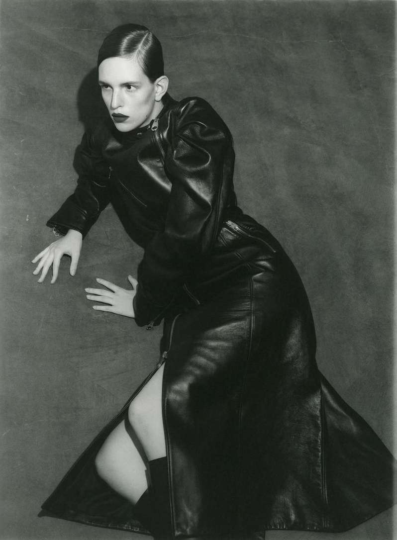 Monochrome studio shot of a fashion model in a long black leather coat with a side slit, lying on the floor with a textured backdrop.