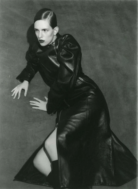 Monochrome studio shot of a fashion model in a long black leather coat with a side slit, lying on the floor with a textured backdrop.