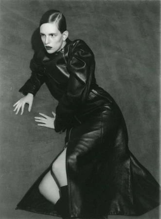 Monochrome studio shot of a fashion model in a long black leather coat with a side slit, lying on the floor with a textured backdrop.
