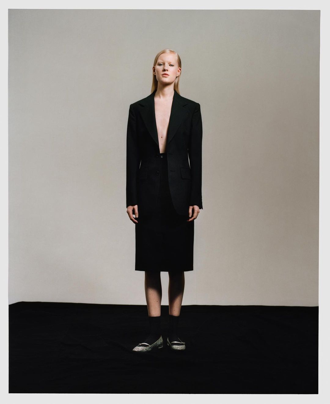 Woman in oversized black suit and skirt stands in a studio setting with muted lighting, conveying a sophisticated and slightly edgy fashion statement.