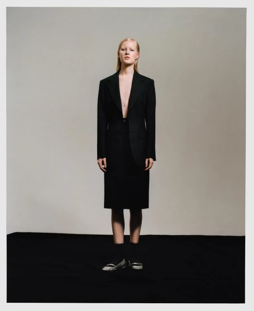 Woman in oversized black suit and skirt stands in a studio setting with muted lighting, conveying a sophisticated and slightly edgy fashion statement.