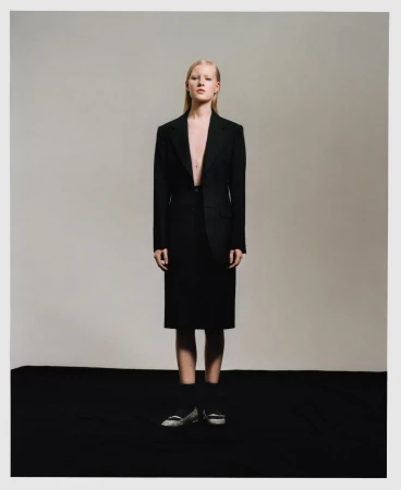 Woman in oversized black suit and skirt stands in a studio setting with muted lighting, conveying a sophisticated and slightly edgy fashion statement.