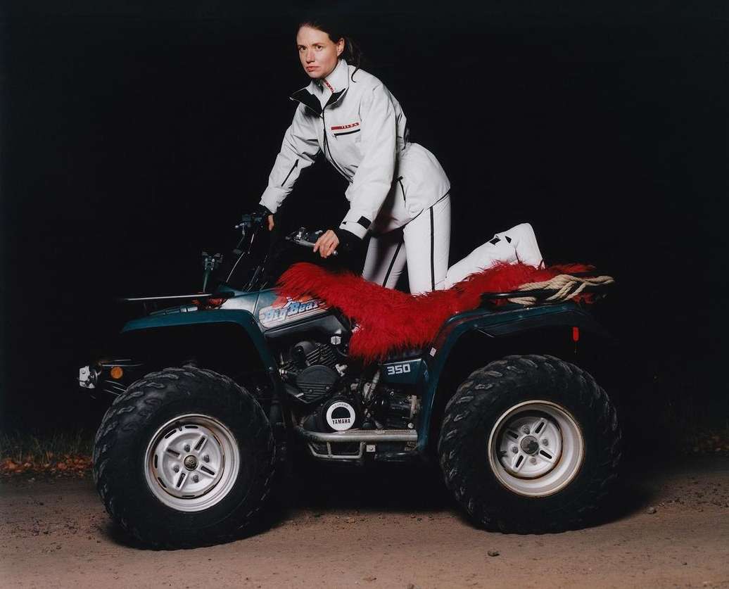 Woman in white suit poses on an ATV with red fur seat cover in a dark outdoor setting, showcasing fashion and outdoor recreation.