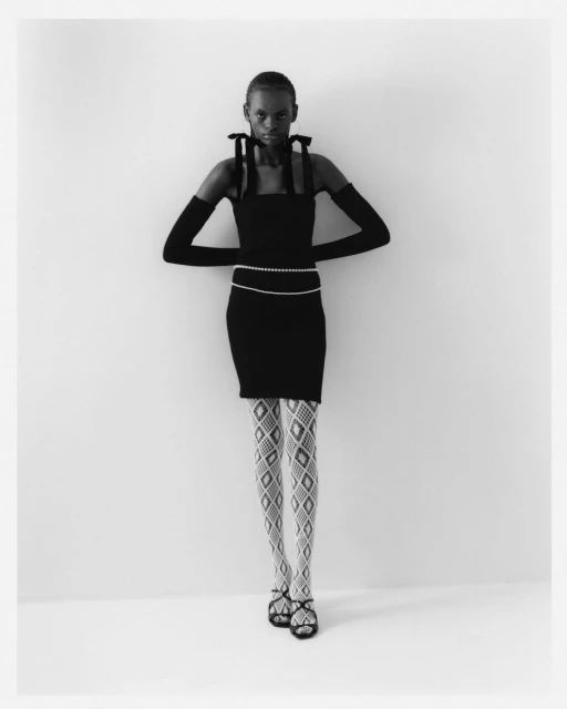 Full-length studio shot of a slender adult woman in a black dress, patterned tights, and sandals against a plain white backdrop.