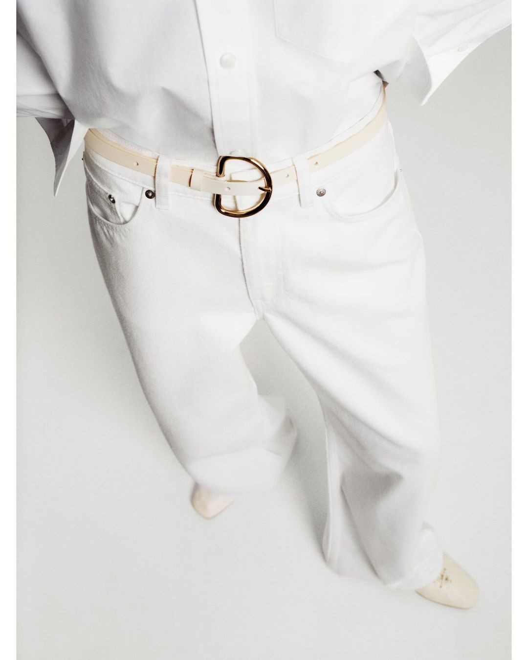 Close-up shot of a person wearing white jeans and a white shirt with a gold-buckled white belt against a white background.