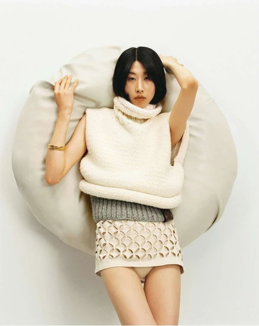 Asian woman in knitted top and skirt lying on beanbag against white backdrop in fashion studio. Posing with hands above head.