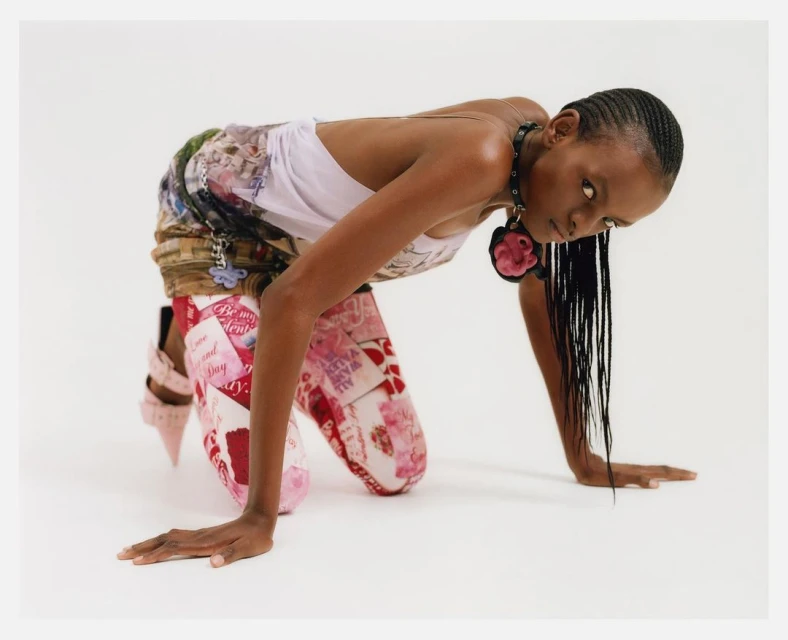Woman in forward-leaning pose, wearing patterned pants and a white top, against a clean backdrop. Braided hair and statement necklace.