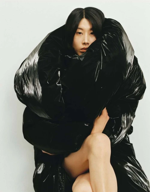 Young woman with dark hair enveloped in an oversized black puffer jacket, against a plain white background in a studio portrait.