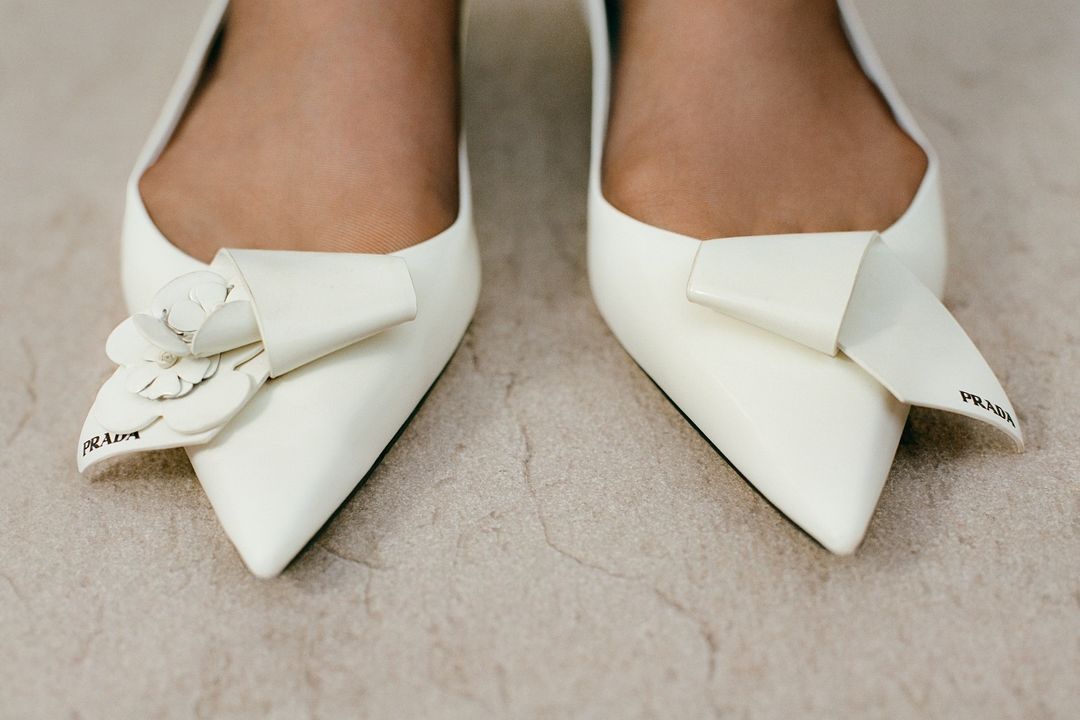 Close-up of pointed-toe white pumps, one adorned with a white flower detail and both featuring Prada logo, on a neutral surface.