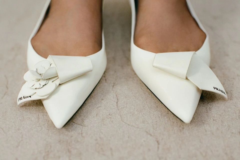Close-up of pointed-toe white pumps, one adorned with a white flower detail and both featuring Prada logo, on a neutral surface.