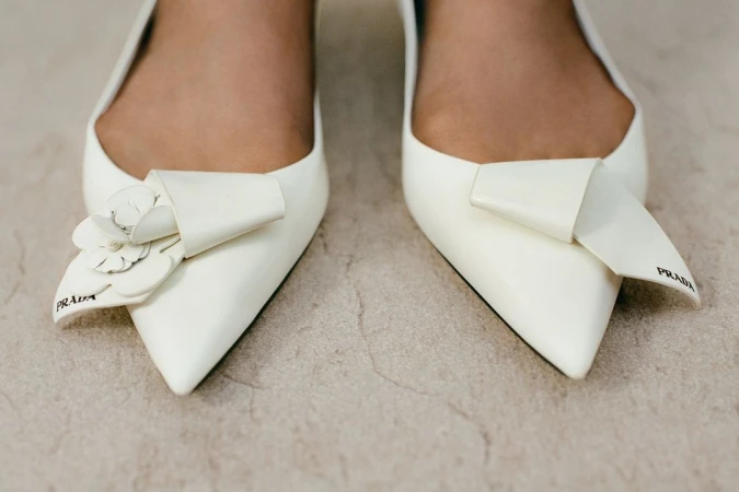 Close-up of pointed-toe white pumps, one adorned with a white flower detail and both featuring Prada logo, on a neutral surface.