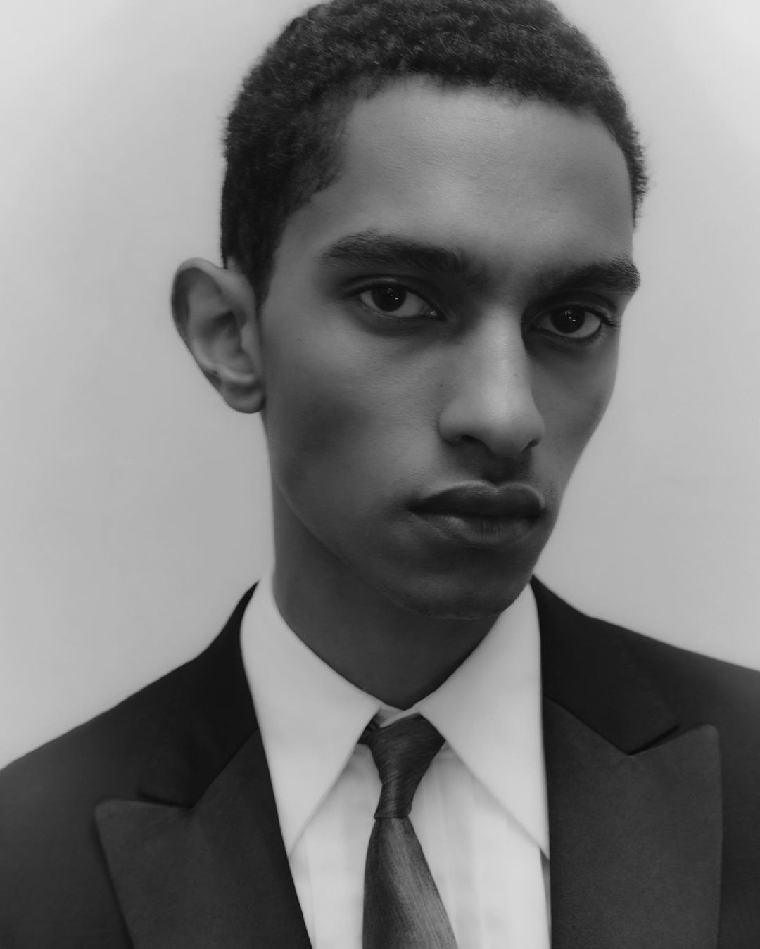 Monochrome portrait of a young adult male in a black suit and tie against a light grey backdrop, close-up head and shoulders.