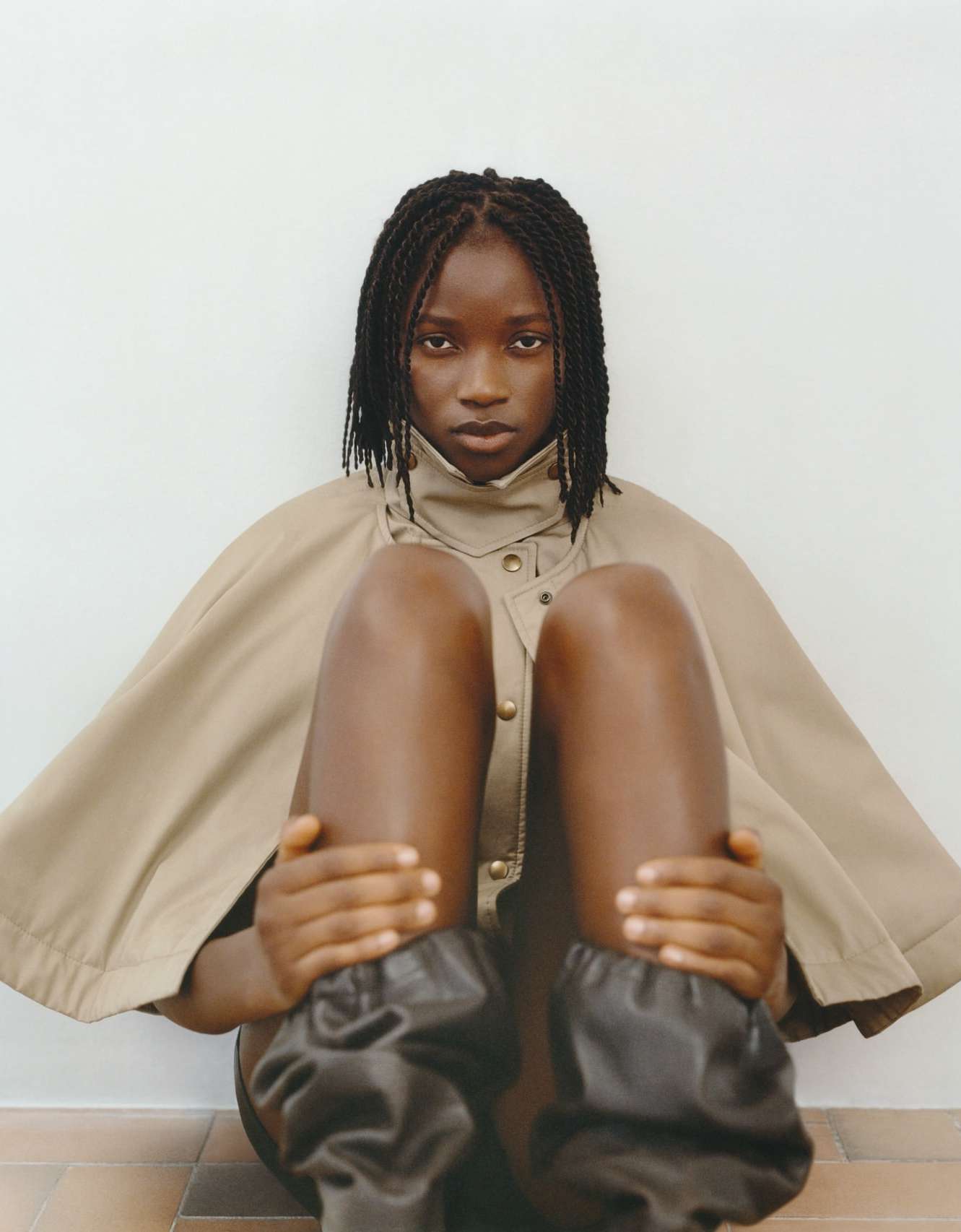 A young Black woman sits indoors wearing a tan cape with oversized black leg coverings, gazing directly at the camera with a serious expression.