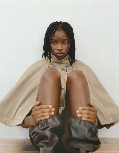A young Black woman sits indoors wearing a tan cape with oversized black leg coverings, gazing directly at the camera with a serious expression.