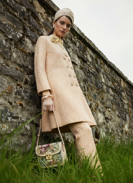 A fashionable woman poses outdoors in a tan double-breasted coat and pants, wearing a matching head wrap and carrying a floral handbag.