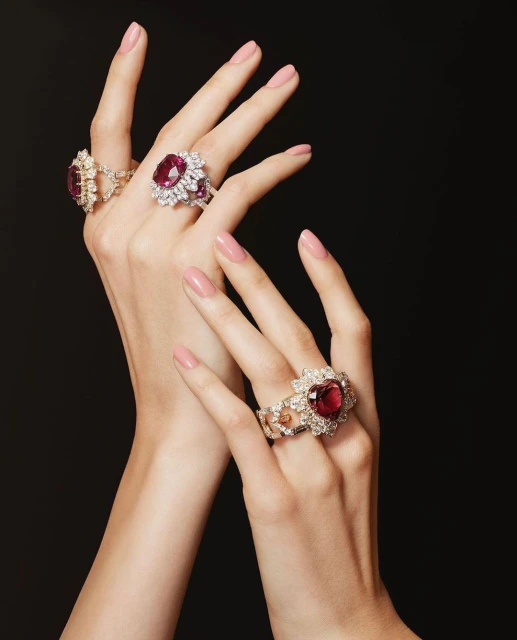 Elegant hands adorned with three diamond rings featuring large ruby gemstones against a stark black backdrop, showcasing beauty and fine jewelry.