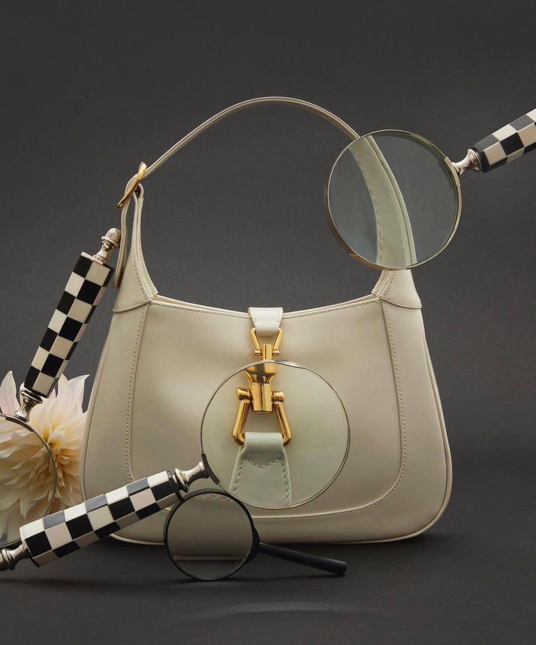 Cream-colored leather purse with gold hardware is displayed with magnifying glasses and a white dahlia against a dark gray background.