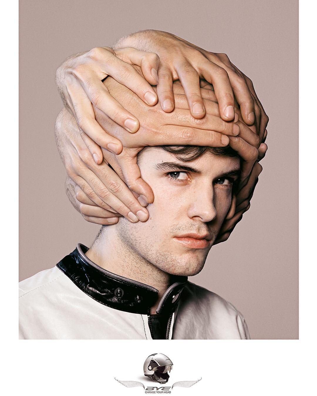 Artistic image of a young man with a 'helmet' made of many hands, wearing a black and white racing jacket, against a taupe background.